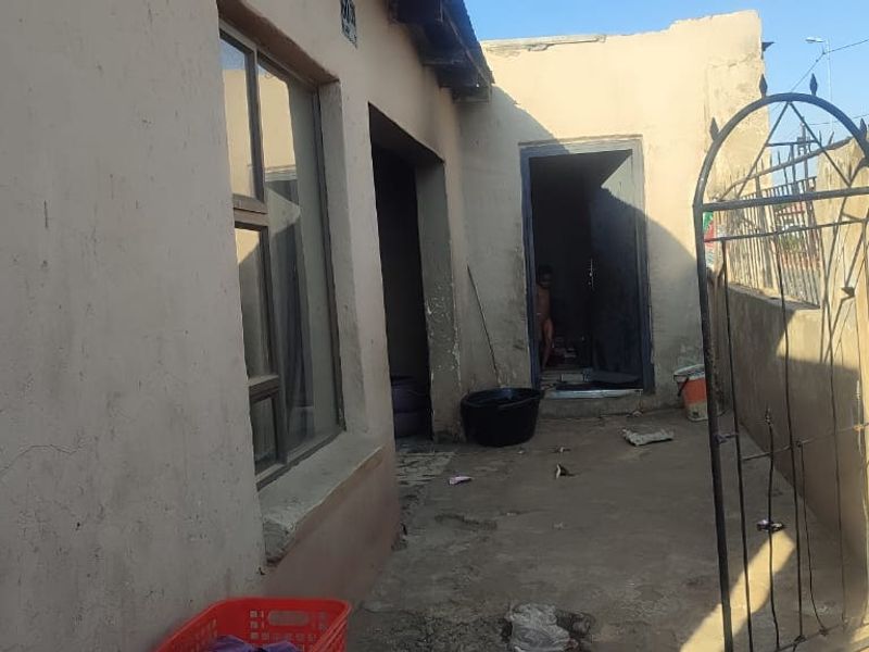 Front exterior view of the property featuring a metal security gate, boundary wall, and basic building facade with visible wear to the plaster finish.