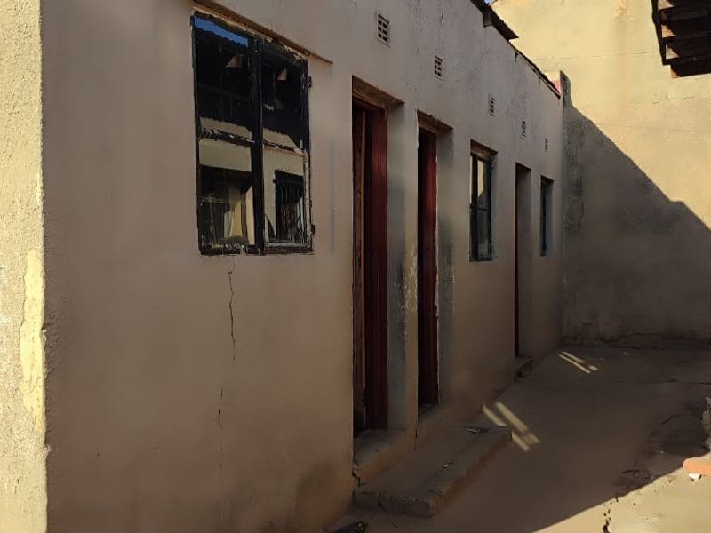 A view of the rear exterior of the property showing plastered masonry walls, multiple access doors, and steel-framed windows in need of maintenance.
