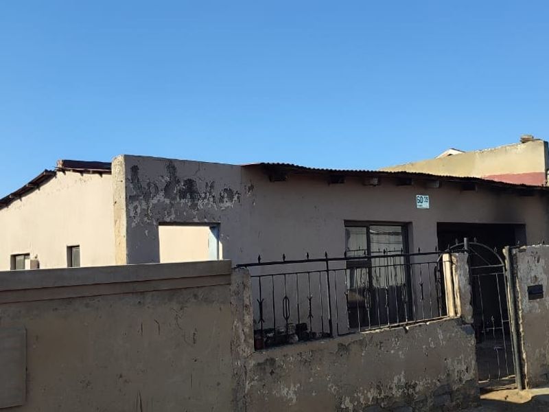 Front exterior view of a single-story house featuring a perimeter wall, metal security gate, and weathered facade requiring maintenance.