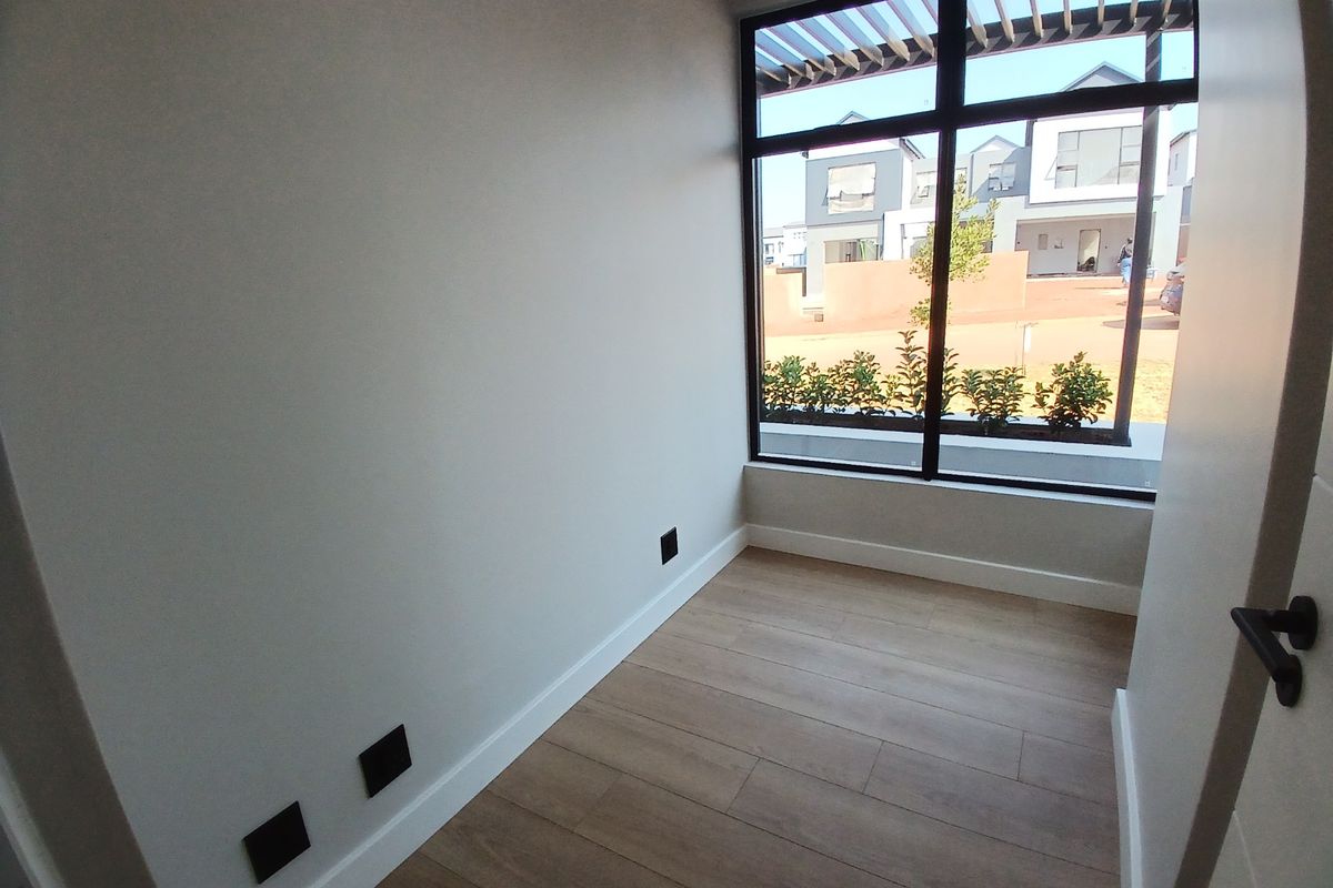 A bright, empty bedroom featuring modern laminate flooring, clean white walls, and a large window providing natural light and a view of the exterior.
