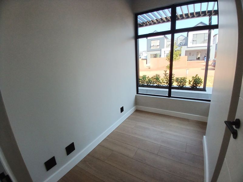 A bright, empty bedroom featuring modern laminate flooring, clean white walls, and a large window providing natural light and a view of the exterior.
