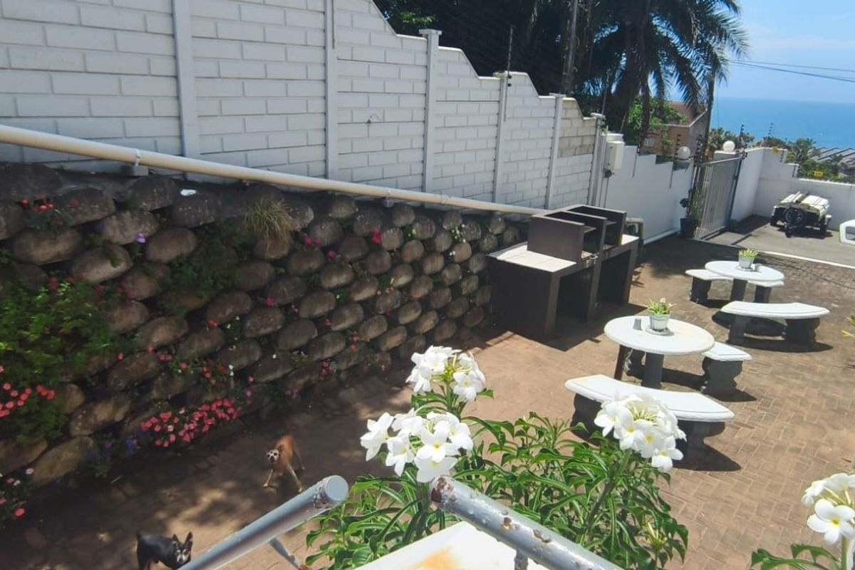 An outdoor rear patio area featuring a built-in braai, stone retaining wall, and paved flooring with views towards the ocean.
