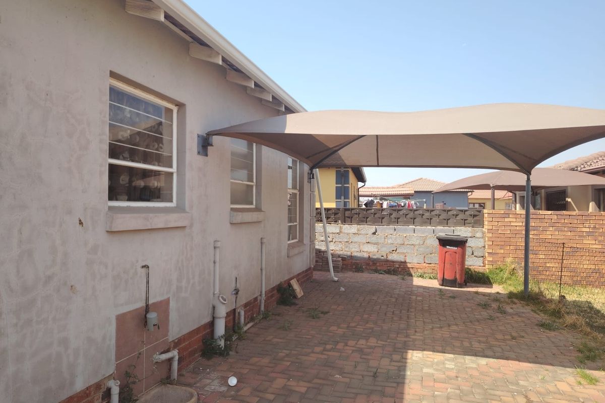 Rear exterior area featuring brick paving, a shade sail structure, and external plumbing fixtures against the house wall.