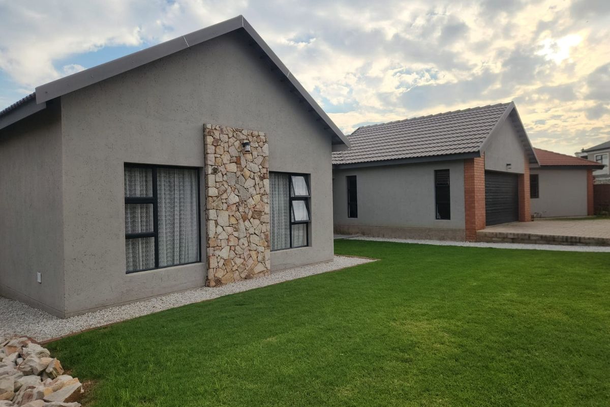 A modern residential exterior featuring stone cladding accents, grey rendered walls, aluminium window frames, and a well-maintained lawn with an adjacent garage.