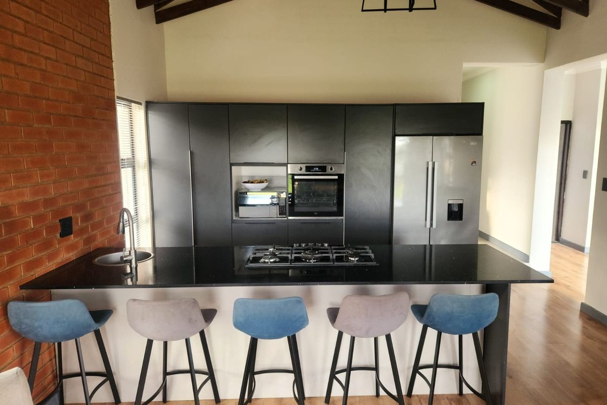 Modern kitchen featuring a large island with gas cooktop, sleek dark cabinetry, built-in oven, and a striking exposed brick wall with timber ceiling beams.