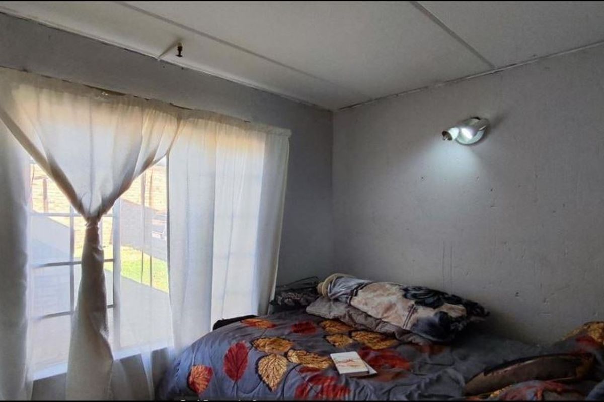 A compact bedroom featuring a window with security bars and a wall-mounted spotlight, requiring cosmetic updates to the walls and ceiling.