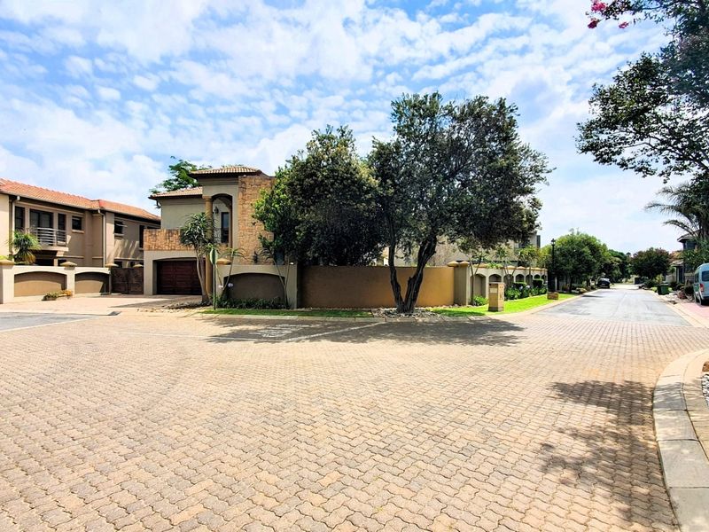 A view of the residential street approach showing the property facade, paved driveway, and surrounding landscaped boundary walls.