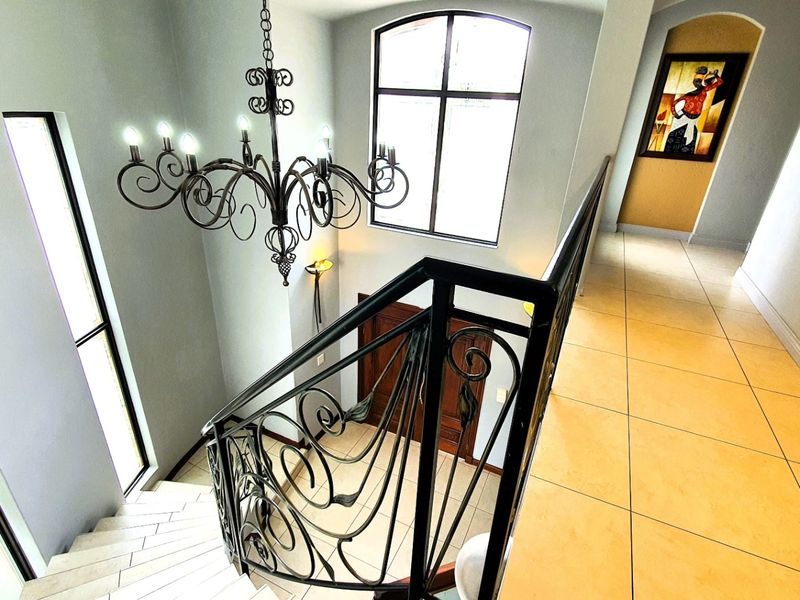 A bright, spacious hallway featuring a decorative wrought iron staircase railing, a matching chandelier, and large windows providing ample natural light.