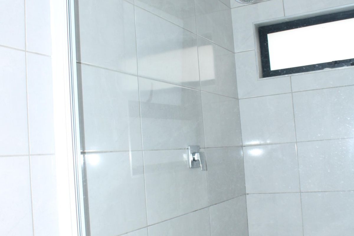 A clean, modern bathroom shower area featuring white wall tiles, a glass enclosure, and a fixed shower head.
