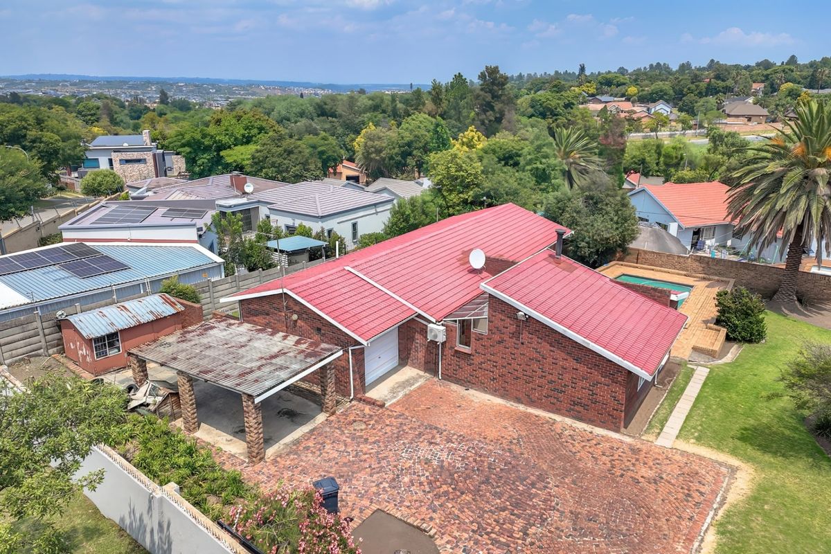 Aerial view of a brick house featuring a red metal roof, paved driveway, carport, and a private swimming pool in a suburban setting.