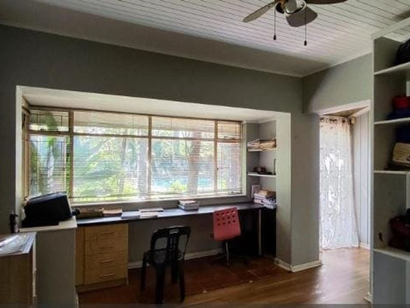 A functional home office featuring a built-in desk, integrated shelving units, timber flooring, and a ceiling fan with large windows overlooking the garden.