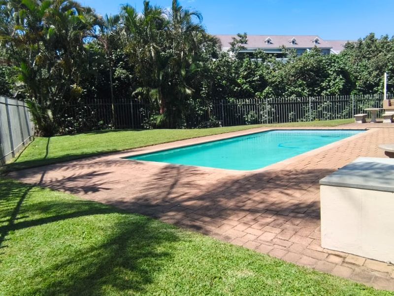 Communal outdoor pool area featuring a paved deck, built-in braai facilities, and well-maintained lawn surrounded by perimeter fencing.