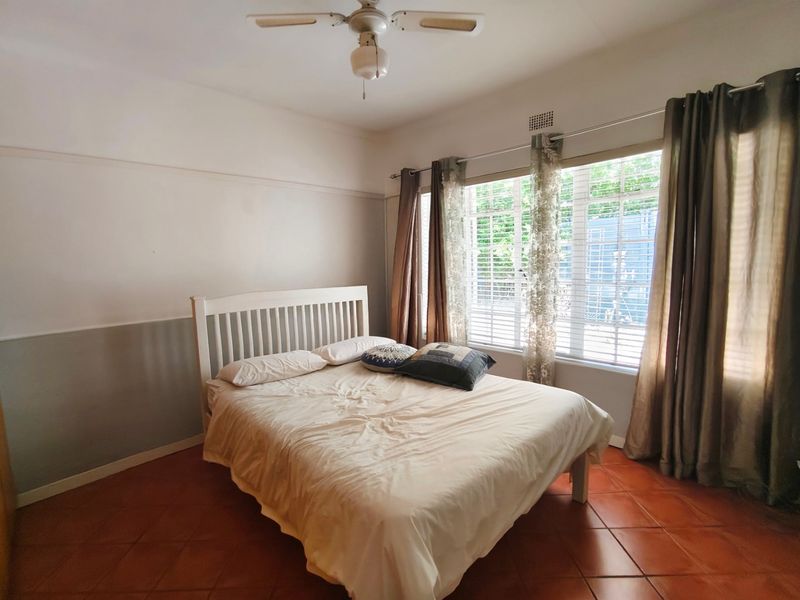 A bedroom featuring terracotta floor tiles, a ceiling fan, and large windows with security bars, offering a functional space with potential for cosmetic updates.