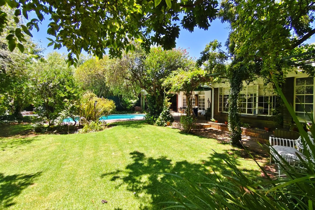 A lush, well-maintained rear garden featuring a swimming pool, mature trees, and a brick patio area adjacent to the house.