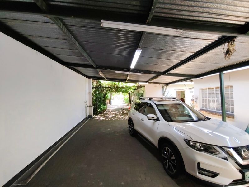 A covered carport area featuring a corrugated metal roof, fluorescent lighting, and paved flooring, providing sheltered parking space.
