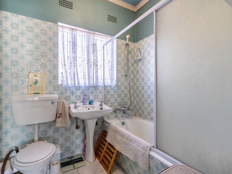 A dated bathroom featuring a pedestal basin, toilet, and a bathtub with a sliding shower screen, finished with vintage patterned wall tiles.