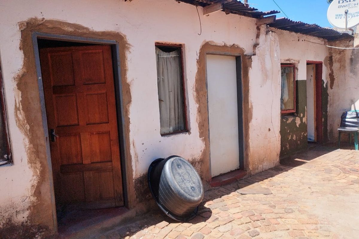 Exterior view of a residential building featuring a corrugated iron roof, cobblestone paving, and multiple entry points with visible wear to the wall finishes.
