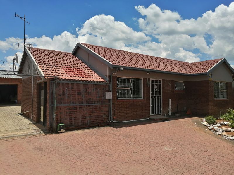 Front exterior view of a single-story brick house featuring a tiled roof, paved driveway, and security installations including bars and razor wire.