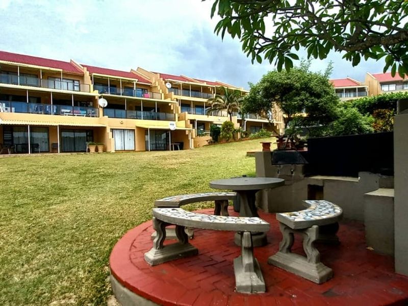 A view of the rear exterior of the apartment complex, featuring a communal lawn and a built-in braai area with paved seating.