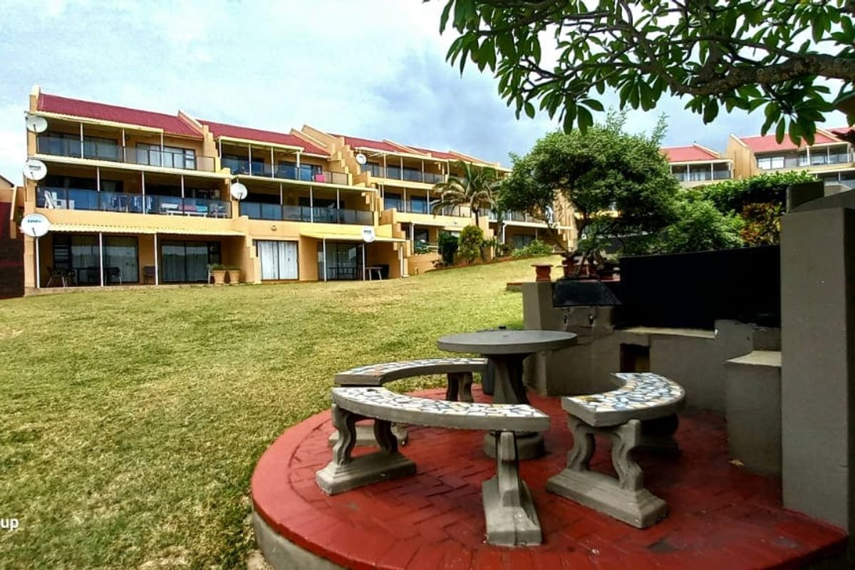 A view of the rear exterior of the apartment complex, featuring a communal lawn and a built-in braai area with paved seating.