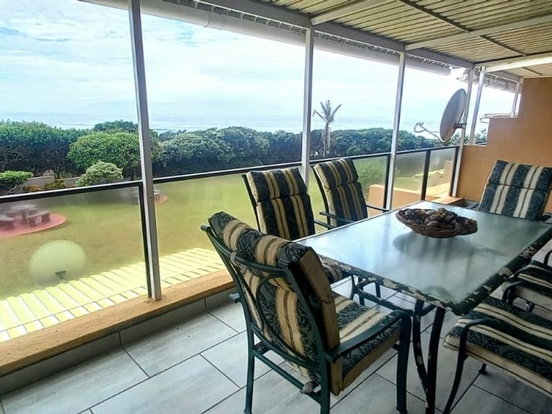 A covered balcony featuring tiled flooring and a glass balustrade, offering scenic views of the garden and ocean beyond.
