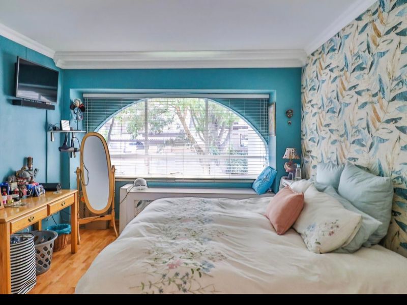 A cozy bedroom featuring hardwood flooring, crown molding, and a distinctive arched window with venetian blinds, accented by decorative wallpaper.