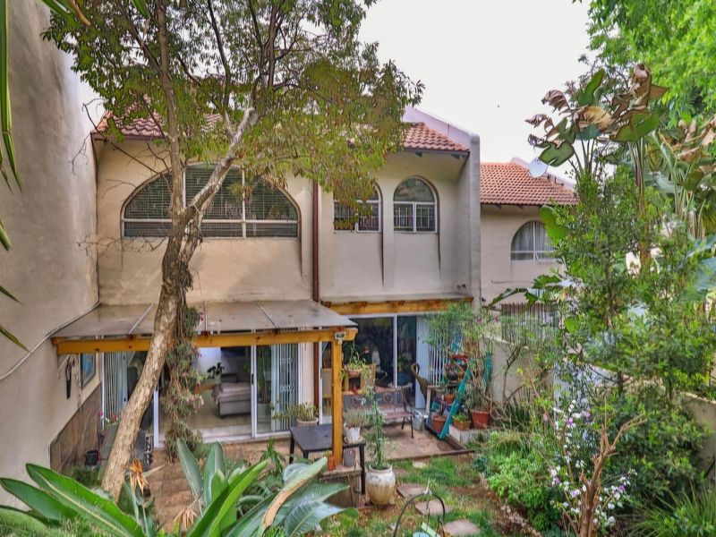 Rear view of a townhouse featuring a paved patio, timber pergola, and arched windows, set within a mature garden area.