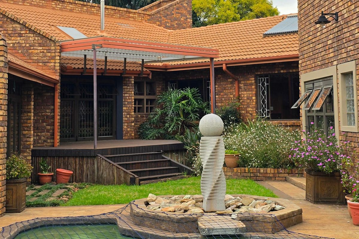 Rear exterior view featuring a facebrick house, timber deck, swimming pool with safety net, and a central stone water feature in the garden.