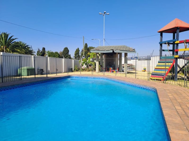 Communal outdoor pool area featuring a paved surround, perimeter fencing, a covered gazebo, and a playground structure.
