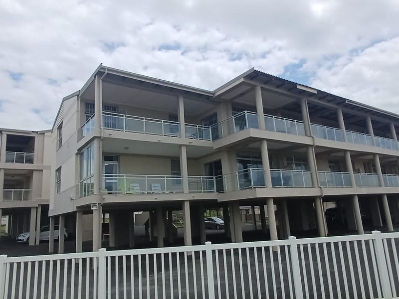A multi-story apartment building featuring elevated construction, covered balconies with glass balustrades, and a paved ground-level parking area enclosed by white fencing.