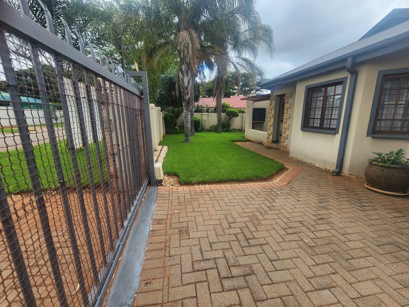 A well-maintained front exterior featuring a paved driveway, secure metal fencing, a neat lawn, and established palm trees.