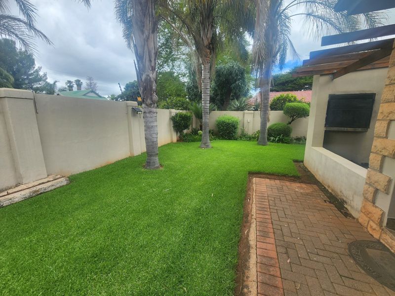 A private rear garden featuring a well-maintained lawn, paved patio area, and a built-in braai under a timber pergola structure.