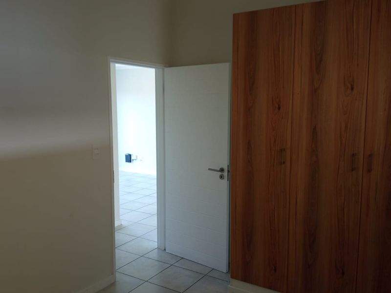 A clean, empty bedroom featuring built-in wooden wardrobes and neutral tiled flooring, with a doorway leading to an adjacent room.