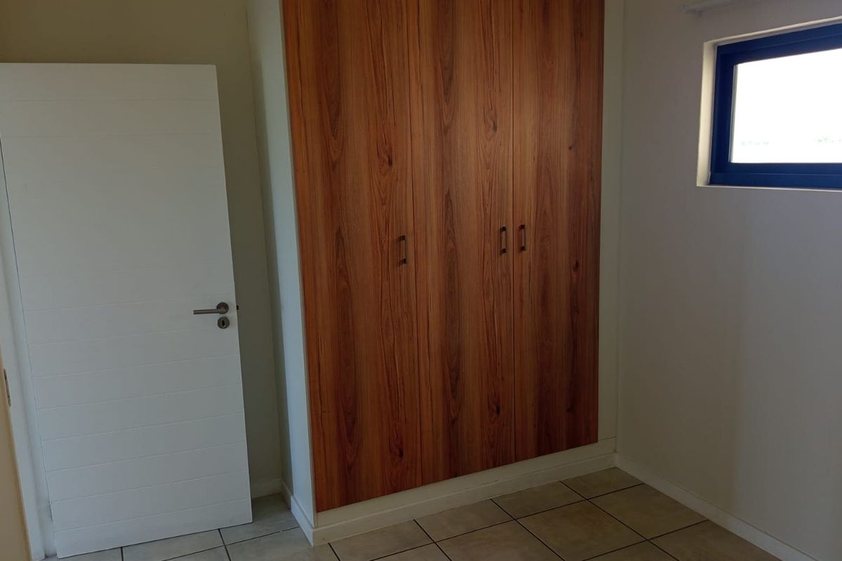 A clean, empty bedroom featuring modern wood-finish built-in wardrobes, neutral tiled flooring, and a sliding window providing natural light.