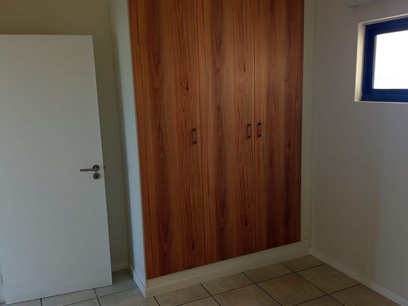 A clean, empty bedroom featuring modern wood-finish built-in wardrobes, neutral tiled flooring, and a sliding window providing natural light.