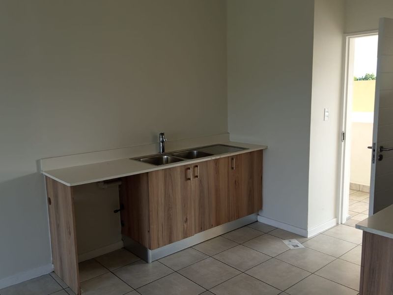 A clean, modern kitchen area featuring wood-finish cabinetry, a double sink, and tiled flooring, with an open layout leading to an outdoor space.