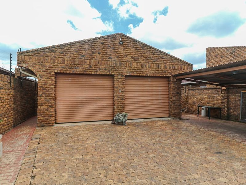 A double garage with face brick exterior, roller doors, and an adjacent carport, set within a paved driveway area with perimeter electric fencing.