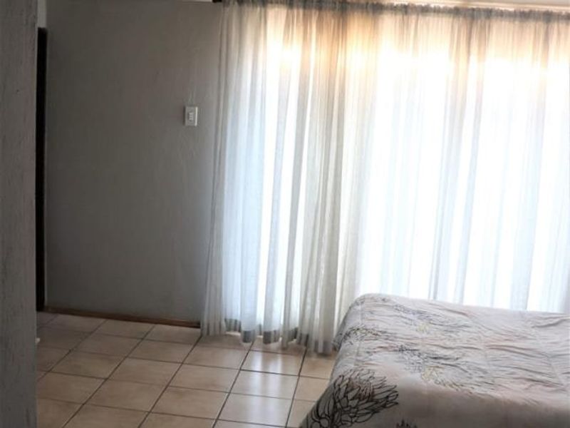 A bedroom featuring tiled flooring and large windows covered by sheer curtains, providing natural light to the space.