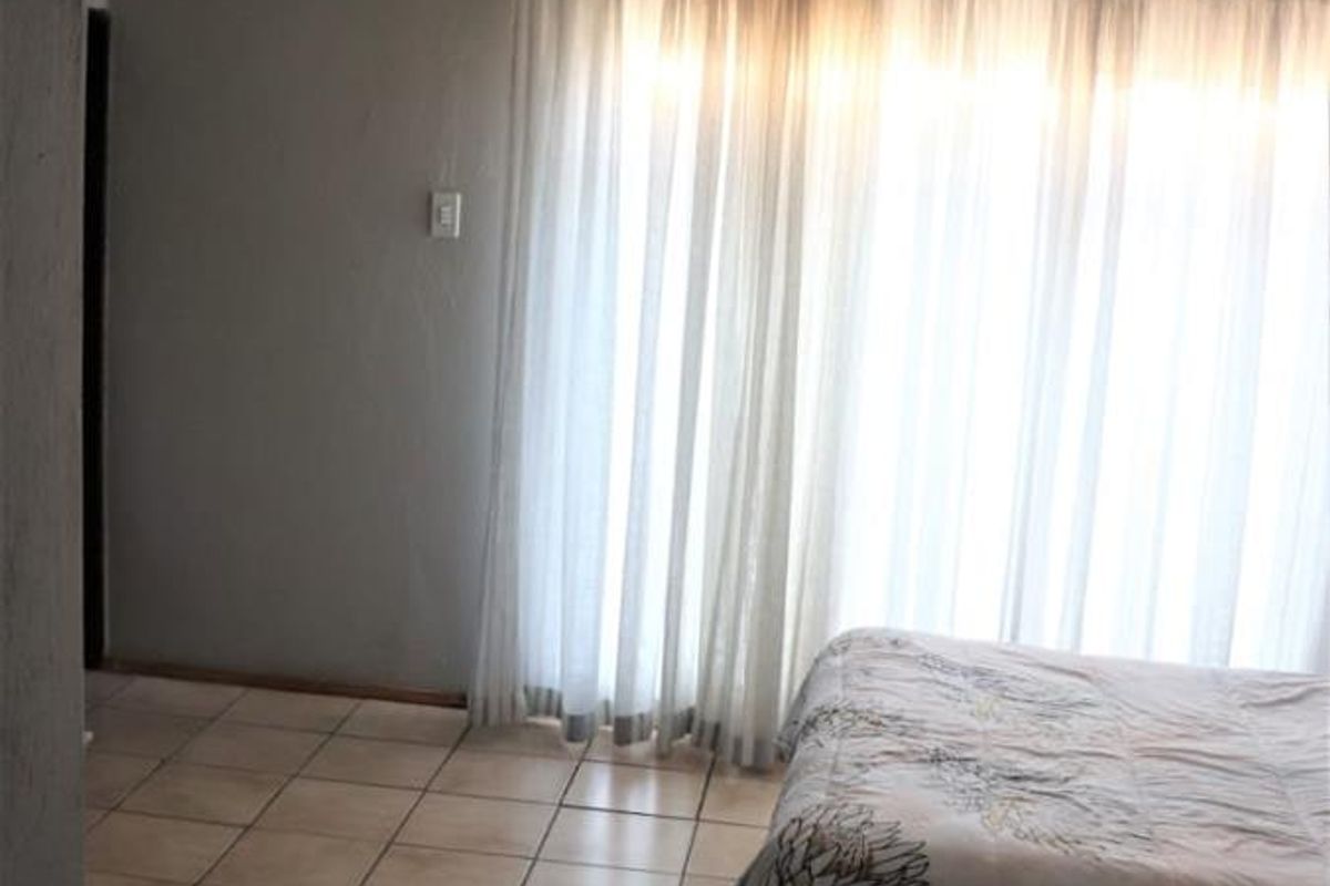 A bedroom featuring tiled flooring and large windows covered by sheer curtains, providing natural light to the space.