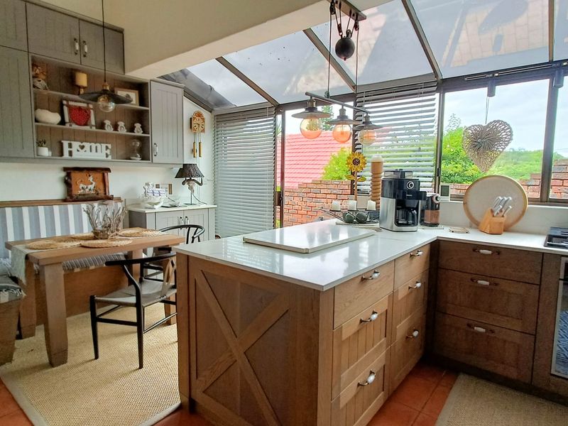 A bright, modern kitchen featuring a central island, stone countertops, built-in cabinetry, and a glass conservatory roof providing ample natural light.