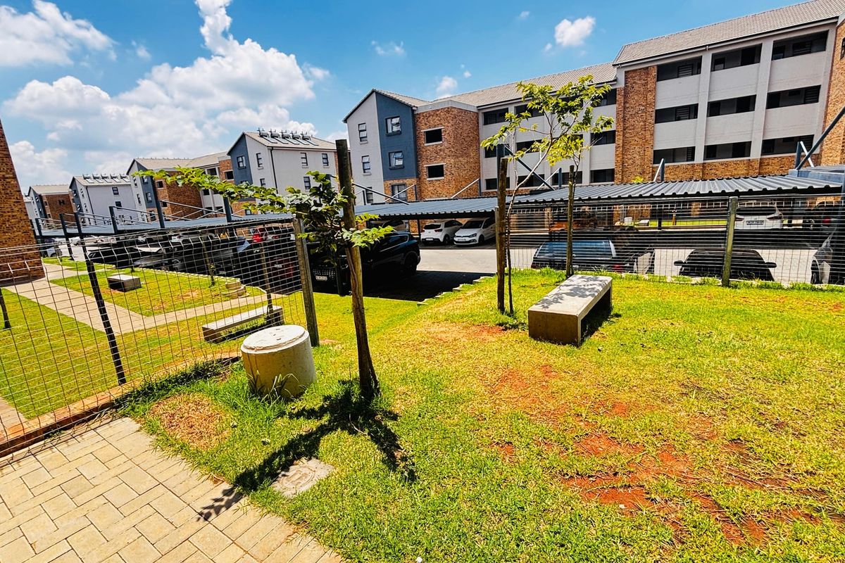 A view of the apartment complex exterior featuring a landscaped garden area, paved walkways, and covered parking bays.