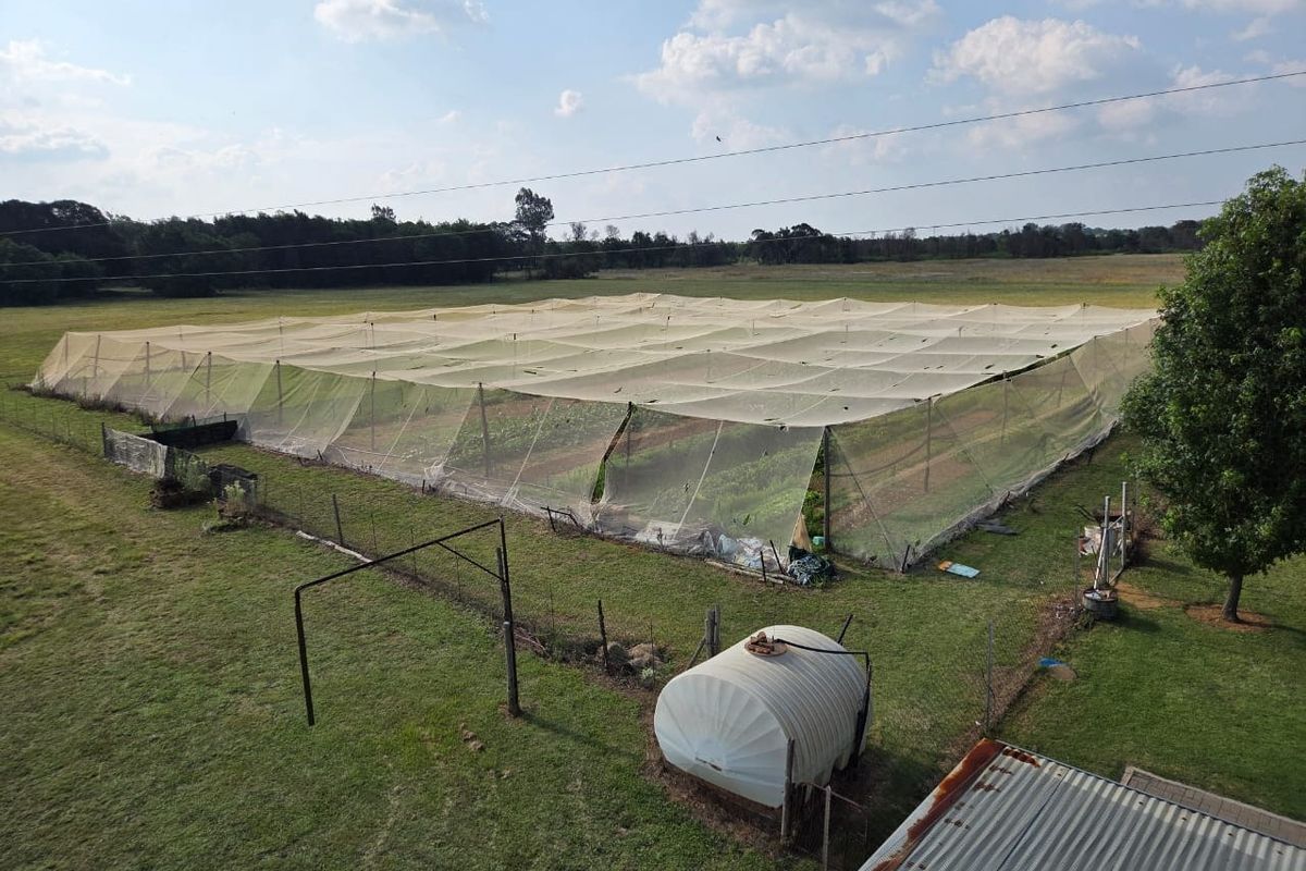 Aerial view of a farm property featuring a large netted agricultural crop enclosure, a water storage tank, and surrounding pasture land.