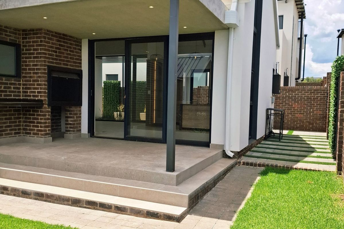 Modern rear exterior featuring a covered patio with a built-in braai, brick chimney, sliding glass doors, and a well-maintained garden.