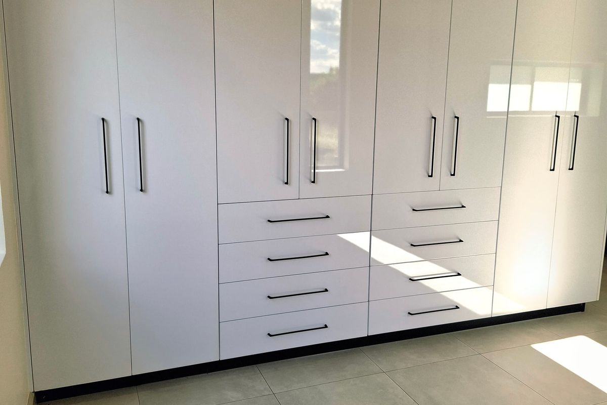 A bright, modern bedroom featuring extensive floor-to-ceiling built-in white cabinetry with integrated drawers and sleek hardware, complemented by neutral large-format floor tiles.
