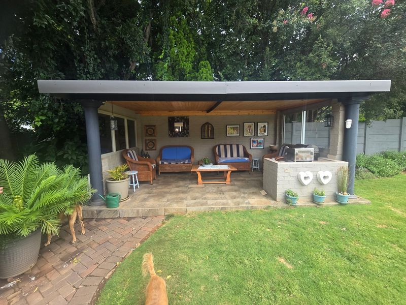 A spacious covered patio featuring a built-in braai, tiled flooring, and a timber ceiling, overlooking a well-maintained garden.