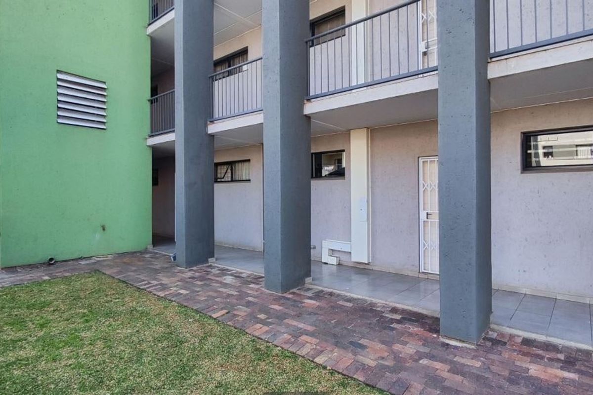 Exterior view of the apartment building featuring a paved walkway, landscaped lawn, and covered patio area with structural columns and security access.