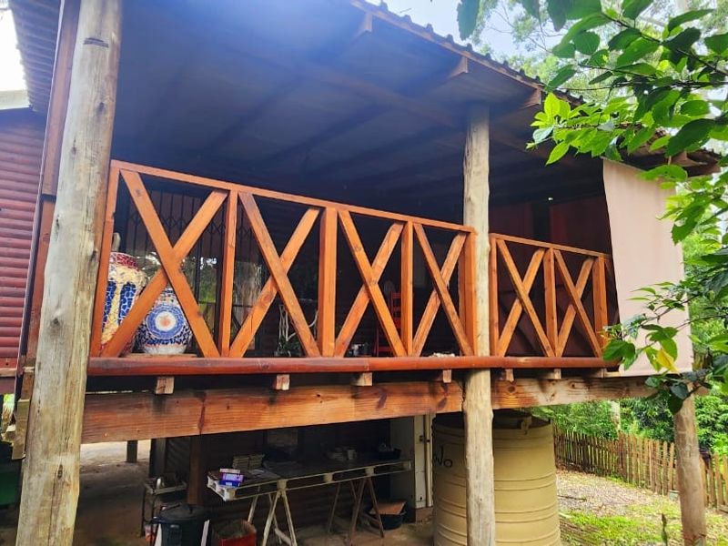A rustic elevated timber balcony featuring a wooden balustrade and covered roof, overlooking a natural garden setting with an integrated water storage tank.