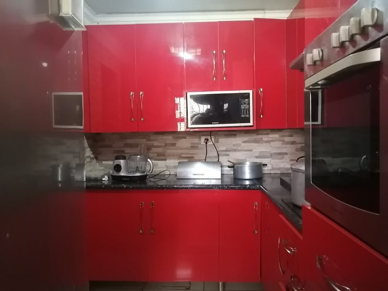 A compact kitchen featuring vibrant red high-gloss cabinetry, dark countertops, a stone-look tile splashback, and integrated built-in oven and microwave appliances.