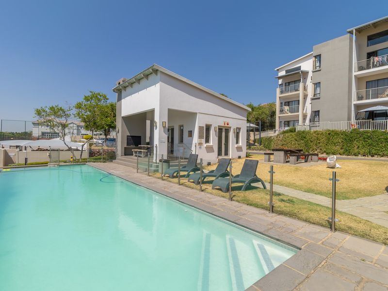 Communal swimming pool area featuring glass fencing, stone paving, and a covered patio with built-in braai facilities within the apartment complex.