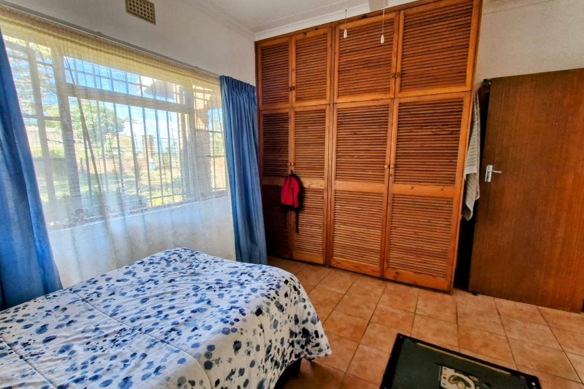 A bedroom featuring built-in wooden louvered cupboards, tiled flooring, and a large window with security bars.
